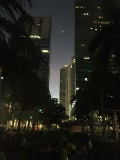 some tall buildings are lit up in the night time sky and people sitting on benches