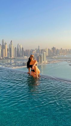 a woman sitting on the edge of a swimming pool in front of a city skyline