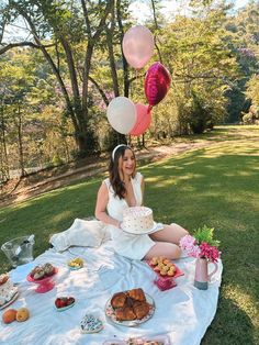 a woman sitting on a blanket with balloons and cake in front of her at a picnic