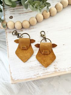 two brown leather cow shaped keychains on a white wooden table with greenery