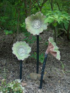 three metal sculptures in the middle of a forest filled with trees and leaves, one has a pink flower on it's head