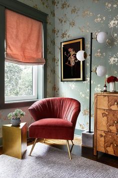 a red chair sitting in front of a window next to a dresser and lamp on top of a rug