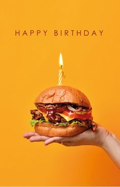 a hand holding a large hamburger with a lit candle in it's middle, against an orange background