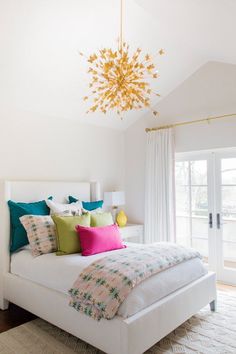 a white bed with colorful pillows in a bedroom next to a door and chandelier