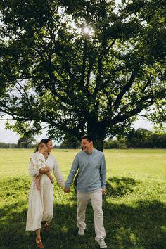 a man and woman holding hands while standing in the grass under a large shade tree