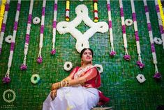 a woman sitting in front of a green wall with flowers and garlands on it