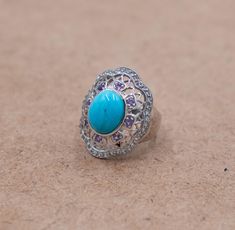 This stunning 9 gram silver flower ring is expertly crafted to dazzle at any event, particularly suited for those who appreciate detailed and vibrant jewelry designs. The centerpiece features an oval turquoise stone, which is encircled by small, sparkling amethyst stones. Adding to its elegance, a line of small sparkling topaz stones outlines the outer rim, enhancing the flower-shaped design on the silver frame. Material: 925 Sterling Silver Weight: 9 grams Center Stone: Oval Turquoise Accent St Silver Turquoise Gemstone Ring For Weddings, Turquoise Ring With Gemstone Accents As Gift, Elegant Silver Turquoise Ring With Gemstone Accents, Silver Turquoise Ring With Gemstone For Wedding, Silver Turquoise Ring For Wedding, Silver Turquoise Ring With Accent Stones For Weddings, Silver Turquoise Wedding Ring With Gemstone, Elegant Handmade Turquoise Wedding Ring, Turquoise Flower Ring Gift