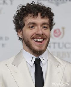 a man with curly hair wearing a white suit and black tie smiling at the camera