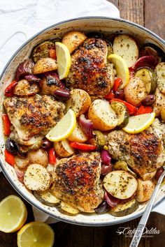 a pan filled with chicken and vegetables on top of a wooden table next to lemon wedges