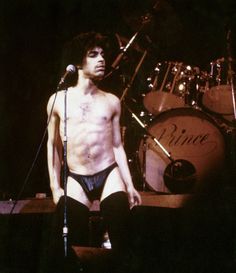 a shirtless man standing in front of a microphone on stage with drums behind him