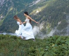 two women in white dresses are running through the grass and trees on a mountain side