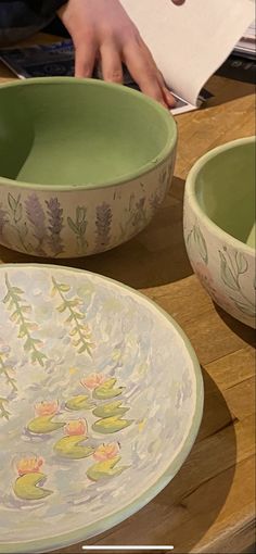 two bowls with flowers painted on them sitting on a table next to a person's hand