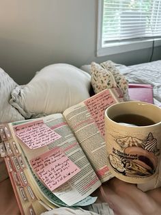 a cup of coffee sitting on top of a bed next to an open book and pillows