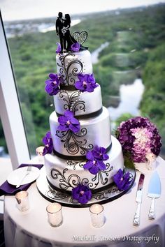 a wedding cake on top of a table with purple flowers in front of a window