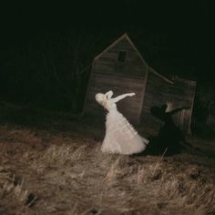 a woman in a white dress standing next to a wooden shed on top of a hill