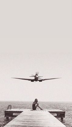 a person sitting on a dock looking at an airplane flying over the ocean in black and white