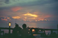 two people sitting on a bench at the end of a pier watching the sun go down