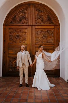 a newly married couple holding hands and standing in front of a wooden door with their veil blowing in the wind