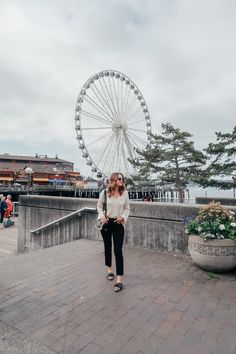 a woman standing in front of a ferris wheel on top of a brick walkway next to water