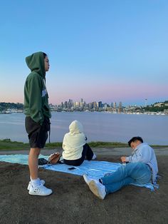 two people sitting on a blanket in front of a body of water with a cityscape in the background