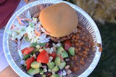 a person holding up a paper plate with a hamburger and salad on it, in front of other food items