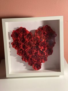 a heart made out of red roses in a white frame on a table next to a pink wall