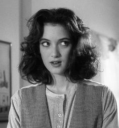 a woman with dark hair wearing a vest and red lipstick looks at the camera while standing in a room