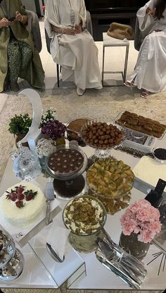 a table filled with cakes and desserts on top of a white table covered in people