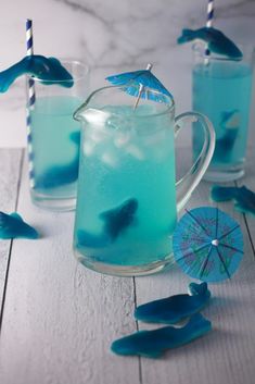 two glasses filled with blue liquid and umbrellas on top of a white wooden table