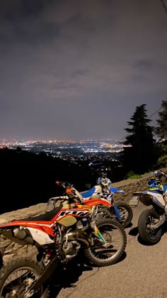 three motorcycles parked on the side of a road at night with city lights in the background