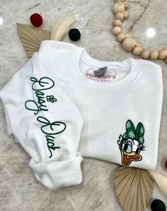 Join the festive fun this holiday season with Mrs. Duck! Wrap yourself in cozy comfort in our soft blended sweater, featuring a jolly Mrs. Duck in a festive Christmas bow embroidered on the chest, and her whimsical autograph along the right sleeve. It's the perfect way to spread holiday cheer with your favorite gal! ✦Sweater design features two embroidered locations. ✦Choose your preferred colored crew! ✦Interested in a different character? Head over to our "All Crewnecks" tab or type 'Christmas Autograph Crewneck' in the search bar to see the other characters! ✦Want to pair this sweater with our mouse ears? Browse our ear selection to find your favorite match! White Tops With Custom Embroidery For Christmas, Green Embroidered Tops For Winter, Holiday Embroidered Casual Sweatshirt, Casual Embroidered Christmas Top, Casual Embroidered Holiday Sweatshirt, Casual Holiday Embroidered Sweatshirt, Casual Festive Sweatshirt For Fall, Casual Fall Festive Sweatshirt, Christmas White Embroidered Tops