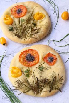 two pizzas with tomatoes and herbs on them sitting on a marble counter top next to green stems