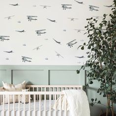 a white crib in front of a wall with airplanes on it and a potted plant