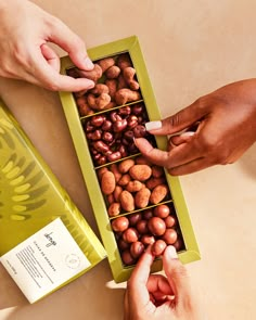 two hands reaching for nuts in a green box on a table next to another person's hand