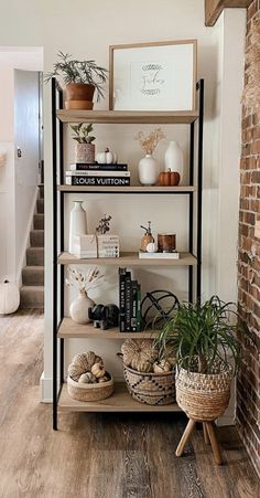 a shelf filled with books and plants in front of a brick wall next to a stair case