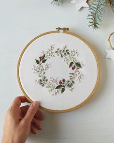 a hand is stitching a christmas wreath on a white wall hanging from a wooden frame