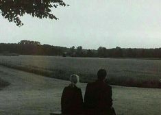 two people are sitting on a bench in the middle of an open field, looking out into the distance