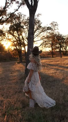 a woman in a white dress and hat standing next to a tree with the sun setting behind her