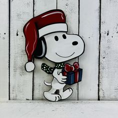 a snoopy christmas decoration with a red hat and bow holding a present on a white wooden background