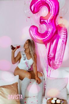 a woman sitting on top of a bed holding a bottle of beer and some balloons