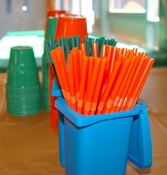 orange and green straws in a blue container on a table next to stacks of plastic cups