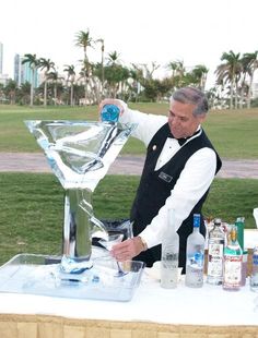 a man pouring drinks into a glass on top of a table in front of palm trees