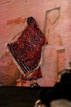 a woman standing on a stage in front of a wall with an egyptian painting behind her