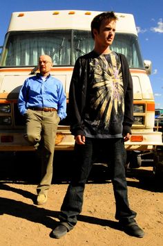 two men standing next to each other in front of a bus on dirt ground with blue sky and clouds behind them