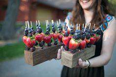 a woman holding a wooden tray filled with berries and strawberries on skewers