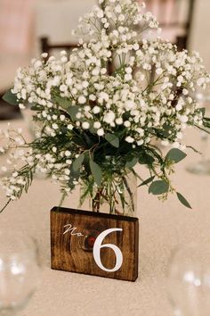 there is a vase with white flowers in it on the table number six, as well as wine glasses