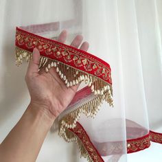 a hand is holding a red and gold garter with white pearls on it, along with sheer curtains