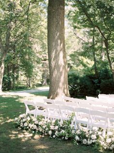 an outdoor ceremony setup with white chairs and flowers on the grass in front of a large tree