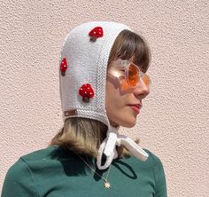 a woman wearing sunglasses and a knitted hat with cherries on the top, standing in front of a pink wall