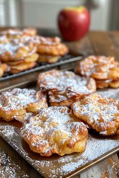 several pastries are on trays with powdered sugar and an apple in the background